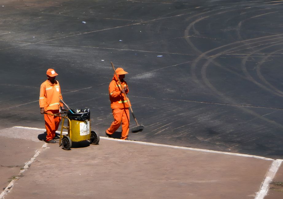 A street cleaning worker dressed in bright orange protective clothing, including a hood and reflective stripes, is sweeping the sidewalk with a long-handled broom. The worker stands next to a green an