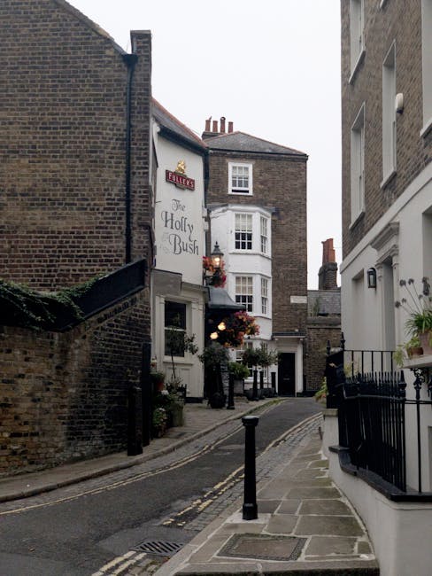 A residential street in Hampstead with a row of Victorian-style terraced houses featuring white facades, bay windows, and brick chimneys under a cloudy sky. The street is paved with asphalt and lined