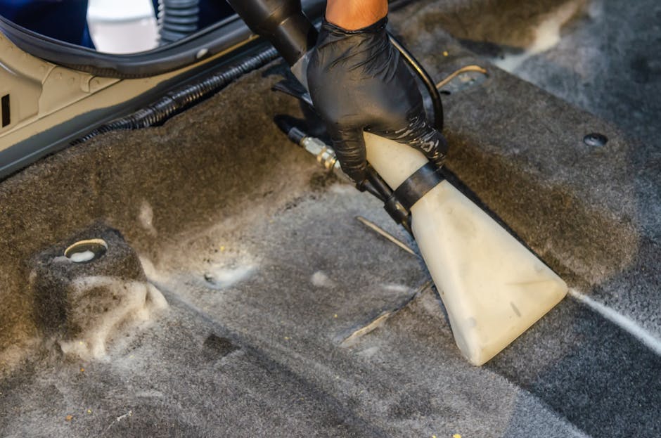 A person kneeling on a patterned area rug in a home setting, preparing a yellow carpet cleaning machine with a black hose attached. The individual, dressed in a beige shirt and blue jeans, is opening